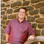 The image shows a man standing against a stone wall with a wheel behind him, smiling at the camera.