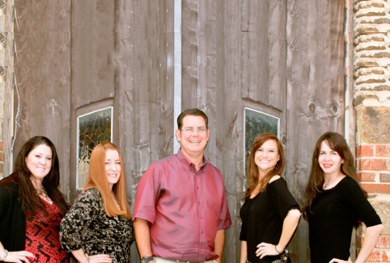 The image features a group of five individuals posing outdoors in front of a rustic wooden structure with a metal door, likely a barn. They are dressed in casual attire, and the setting appears to be during daylight hours.