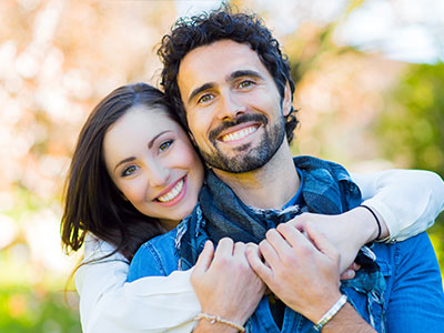 The image shows a man and a woman embracing each other with smiles, set against an outdoor background.