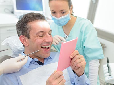 The image shows a man sitting in front of two dental professionals, holding up a pink card with a surprised expression on his face, while they appear to be examining him with dental instruments.