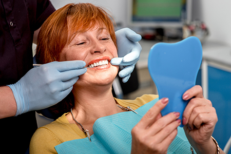 A woman with red hair sits in a dental chair, smiling, while holding up a blue toothpaste tube and looking at her reflection in a mirror.