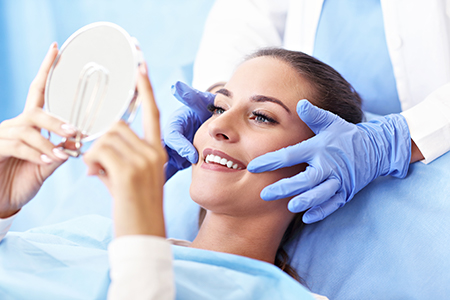 A woman receiving a facial treatment with a device held by a person wearing blue gloves, against a backdrop of a medical setting with a mirror reflecting their face.