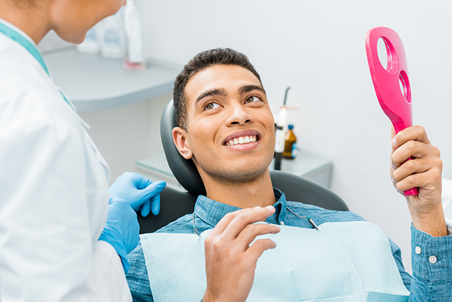 A man in a dental chair is smiling while holding a pink object resembling a toothbrush, with a dental hygienist standing behind him, both in a dental office setting.