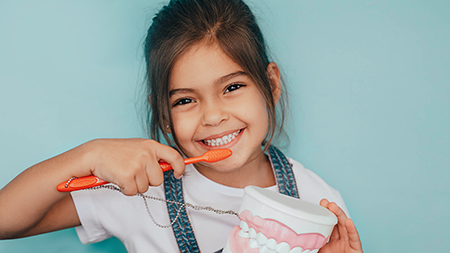 The image shows a young girl brushing her teeth with a red toothbrush while holding a toothpaste tube, smiling at the camera, and looking directly into it.
