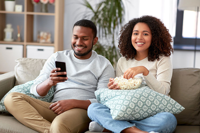 Man and woman sitting on couch, smiling at each other while holding snacks, with a cell phone between them.