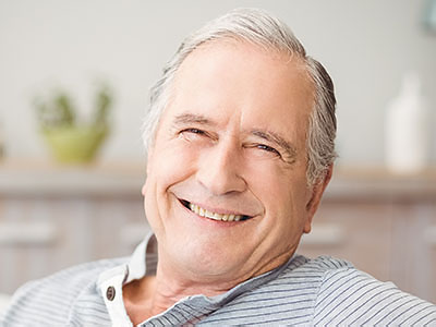 An elderly man with gray hair is smiling broadly, looking directly at the camera while seated in a comfortable chair.