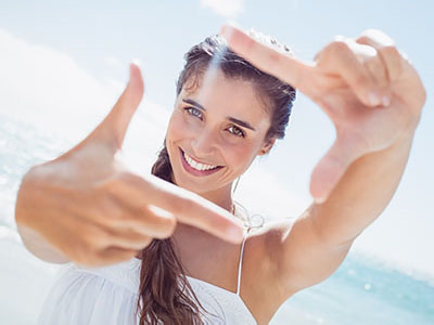 The image shows a woman taking a selfie with her hands framing the camera lens, against a bright background with clear skies.